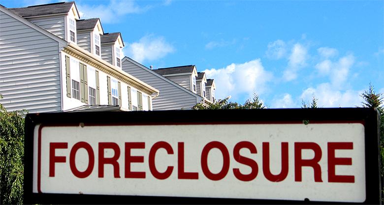 The image shows a 'Foreclosure' sign in the foreground and the tops of a couple of houses in the background.