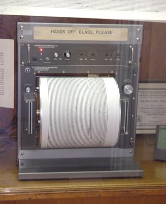 The figure shows a seismograph put on a wooden table. Its top is labeled as 'Hands off glass, please'. Below it there are some buttons are shown and a paper roller is fitted in the seismograph to print the observation by the machine. On the right and left of the roller, two vertical cable slots are given.