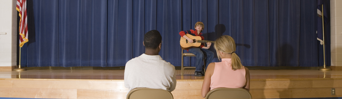 Image of boy with guitar performing on stage in front of two adults in the audience
