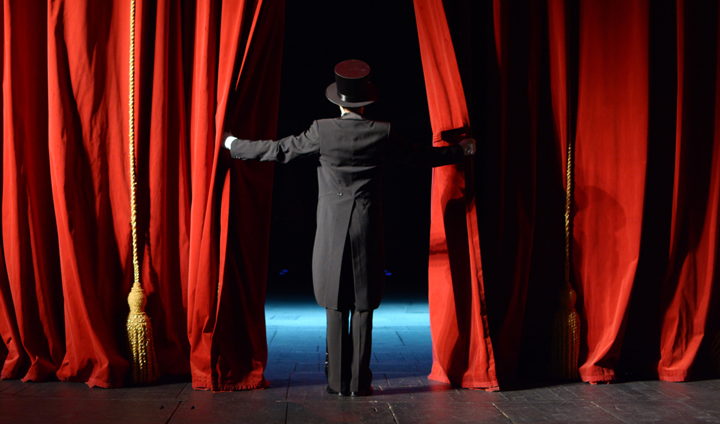Image of child in costume on stage looking out into the audience from behind a red curtain